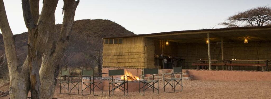 Chairs around a fire pit at dusk, with a thatched dining boma and hillside beyond.
