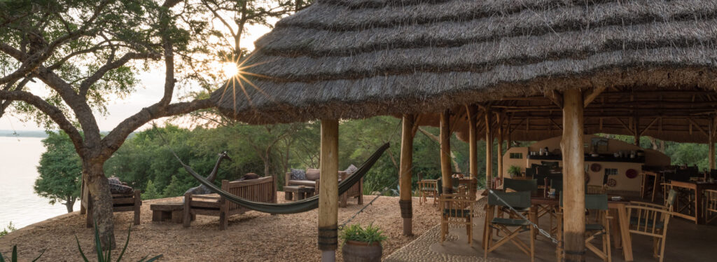 A thatched open sided restaurant with dining tables and a hammock, set on a lakeside deck shaded by a large tree at sunset.
