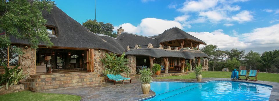 A stone and thatch lodge with a covered veranda, fronted by a curved swimming pool and stone patio with loungers, under a partly clouded sky.