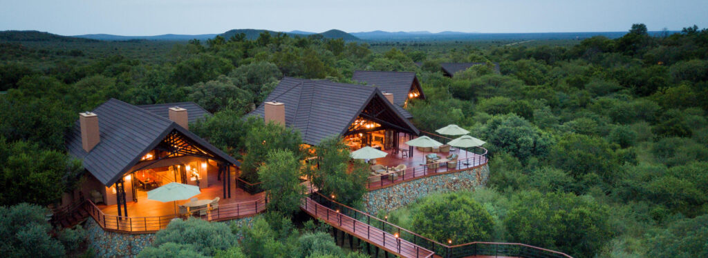 Aerial view of a lit lodge with pitched roofs and a raised walkway leading to a circular fire pit deck, set amid dense bushveld at dusk.