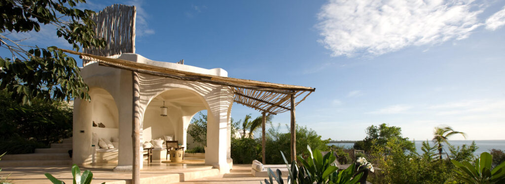 A white domed pavilion with arched openings and lounge seating beside a small pool, framed by frangipani and overlooking the ocean.