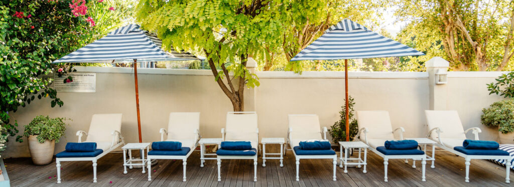 A row of white loungers with navy cushions on a wooden pool deck, shaded by striped umbrellas and a flowering tree.