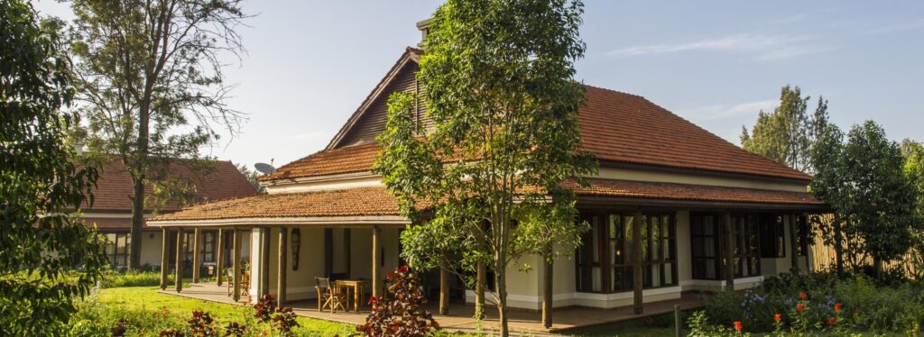 A colonial style farmhouse in Tanzania with a red tiled roof and a covered veranda with wooden columns, set among trees and flowering gardens. legendary lodge.