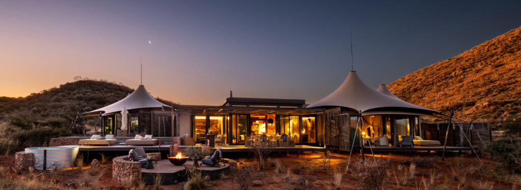 A tented lodge home with tensile canvas roofs, lit from within, with a deck, fire pit and plunge pool set against desert hills at dusk.