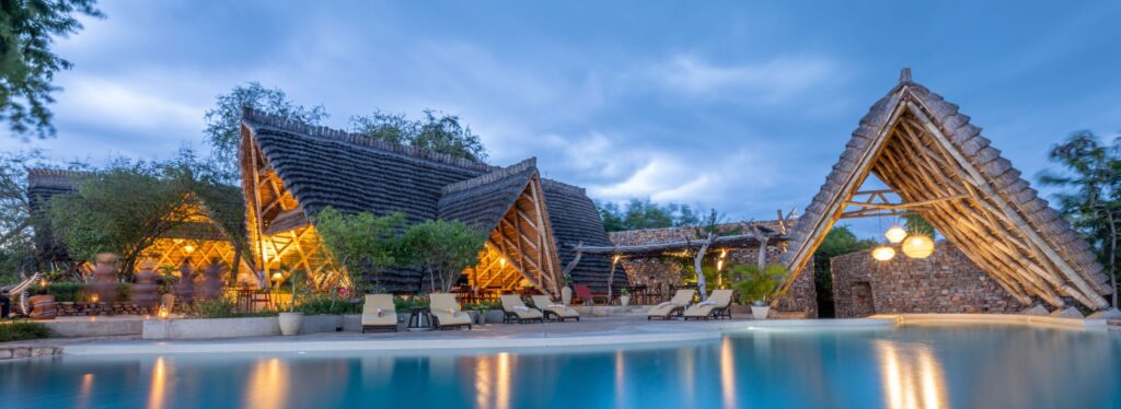 A swimming pool lined with loungers beside thatched, angular roofed buildings lit from within at dusk, with a stone archway nearby.