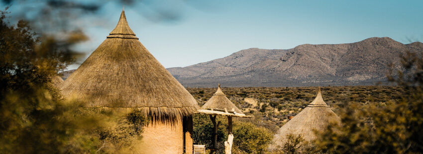 Thatched conical huts seen through bushveld, with hills in the distance.