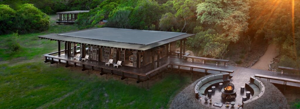 Aerial view of a raised lodge and circular fire pit deck, surrounded by dense forest at sunset.