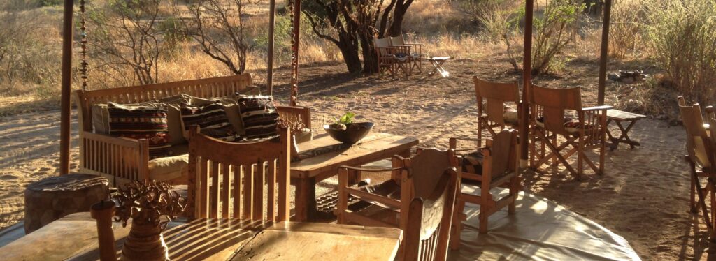 A safari mess tent with wooden benches, director's chairs and patterned cushions, opening onto dry savannah bushland in late afternoon light.
