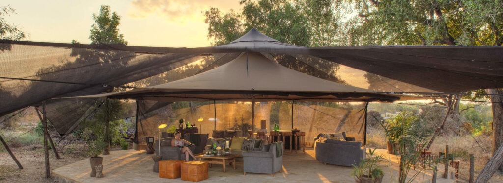 A canopied lounge and bar area with grey sofas and armchairs on a stone terrace, where a guest reads at sunset beneath tall trees.