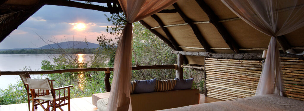 A canopied bed and open sided lounge area beneath a thatched roof, overlooking the Rufiji River at sunset.
