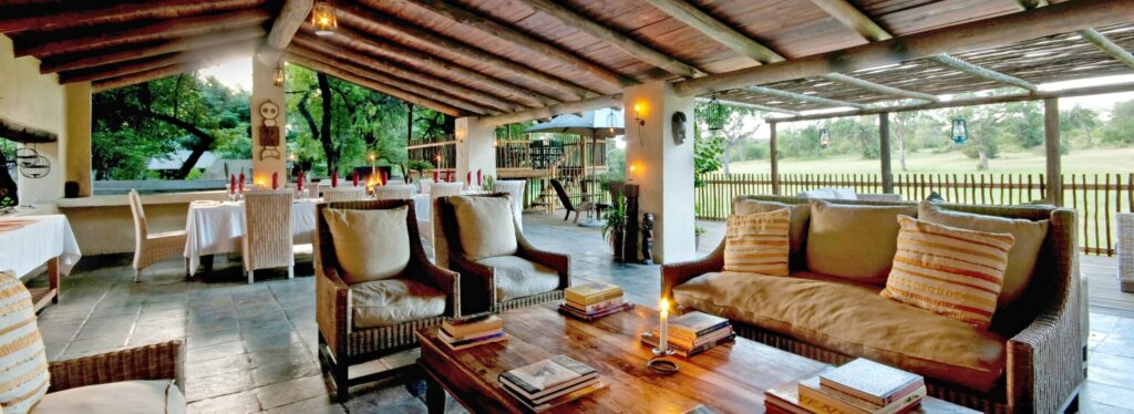 A thatched lounge with wicker sofas and armchairs beside a candlelit dining table, opening onto a deck overlooking open bushveld at dusk.