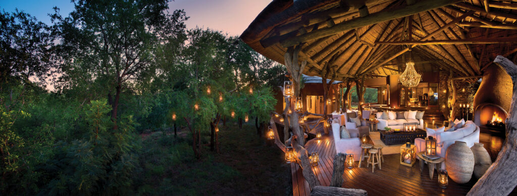 A thatched lounge with white sofas and a chandelier, opening onto a lantern-lit deck strung through trees at dusk.
