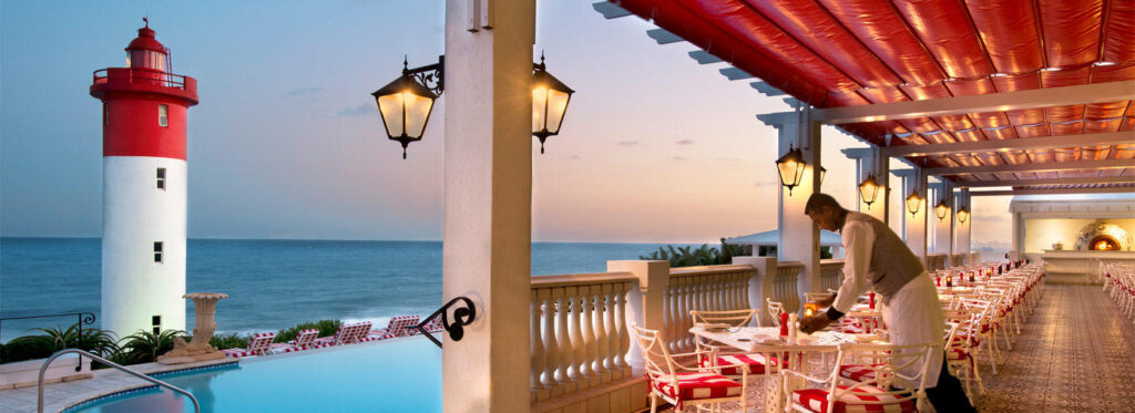 A waiter attends a table on a covered terrace beside a pool, with a lighthouse and sea at dusk.