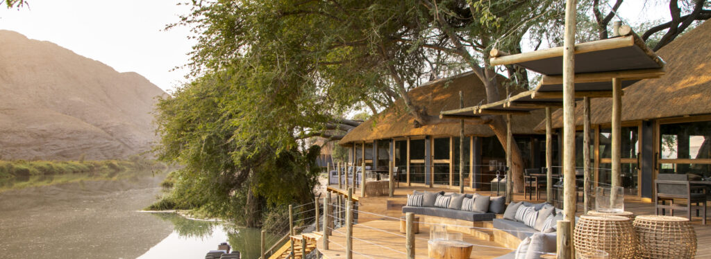 Riverside wooden deck with lounge seating and a thatched lodge, overlooking a river with desert hills beyond.
