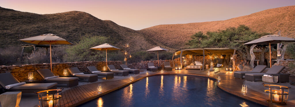 A lantern-lit pool terrace with loungers and umbrellas, enclosed by a stone wall with hills beyond.